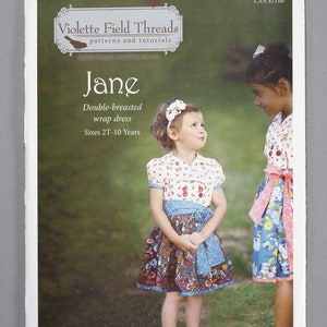 May include: A sewing pattern for a double-breasted wrap dress called "Jane" from Violette Field Threads. The pattern is for sizes 2T-10 years. The image shows two girls wearing the dress in a floral print.