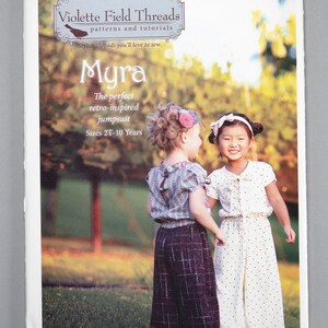 May include: A sewing pattern for a children's jumpsuit called "Myra" by Violette Field Threads. The pattern is for sizes 2T-10 years and features a retro-inspired design. The pattern cover shows two girls wearing the jumpsuit in a garden setting.