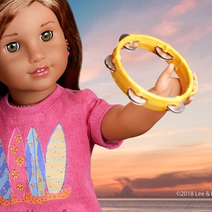 May include: A doll with long brown hair wearing a pink shirt with a surfboard design holds a yellow tambourine. The background is a sunset over the ocean.