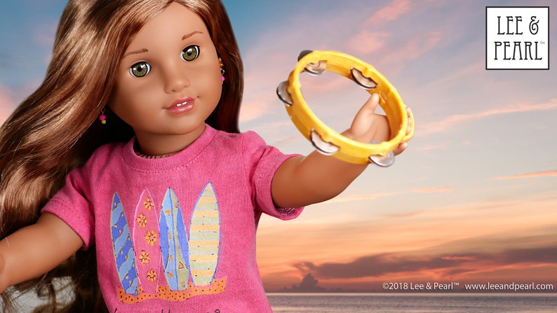 May include: A doll with long brown hair wearing a pink shirt with a surfboard design holds a yellow tambourine. The background is a sunset over the ocean.
