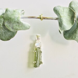 May include: A silver pendant featuring a green aventurine crystal and a moonstone. The pendant is set against a white background with a eucalyptus branch.