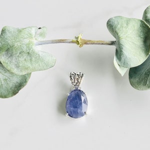 Puede incluir: Un colgante de plata con una piedra preciosa grande, ovalada, de color azul oscuro con corte facetado. El colgante tiene una fianza de plata delicada e intrincada.