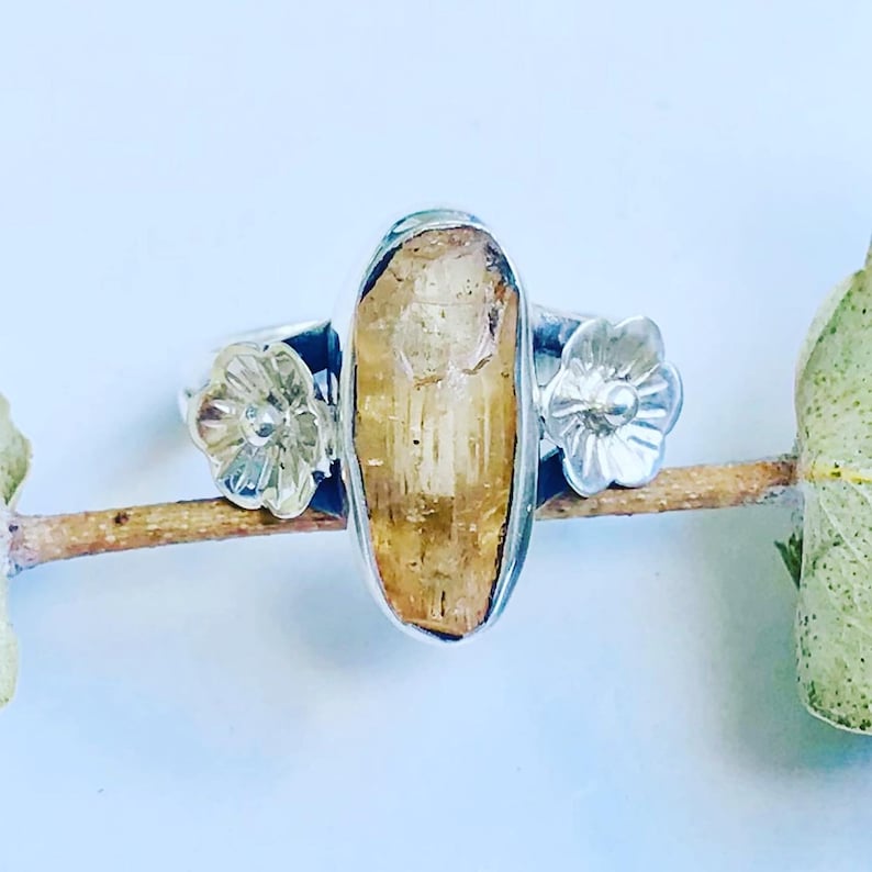 May include: A silver ring with a large, oval-shaped, yellow citrine gemstone. The ring has two silver flower accents on either side of the gemstone.