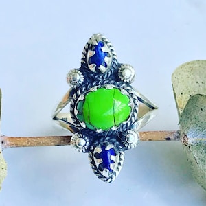 May include: A silver ring with a green stone and blue accents. The ring has a detailed design with a rope-like pattern.