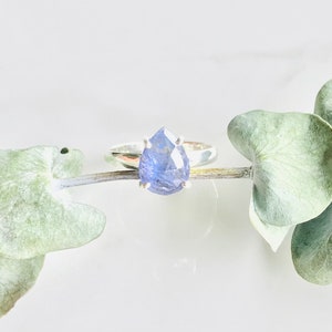 May include: A silver ring with a pear-shaped blue gemstone set in a prong setting. The ring is resting on a branch with green leaves.
