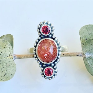 May include: A silver ring with a large, round, orange sunstone and two smaller, round, red gemstones. The ring is set in a beaded design.