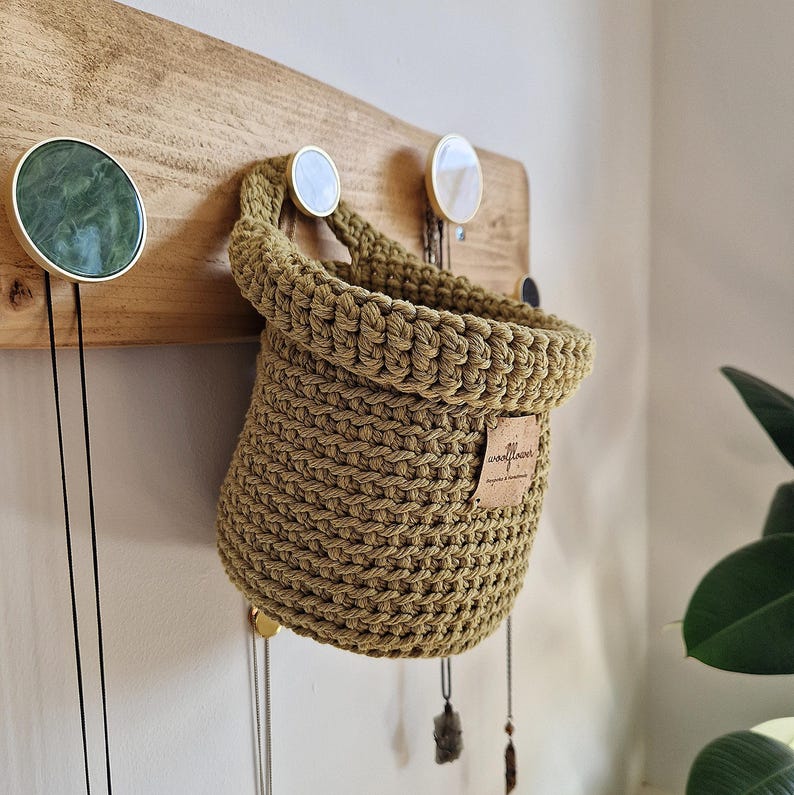 A crocheted hanging basket in a muted green colour with a small brown vegan leather tag that says "woolflower". The basket is hanging on a wooden wall with gold hooks.