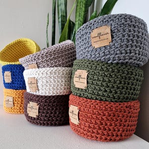 May include: A collection of round, crocheted storage baskets in various colours including yellow, blue, orange, white, brown, grey, and olive green. Each basket features a square cork label with the text woolflower printed in black ink.
