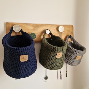 May include: Three hanging crochet baskets in navy blue, olive green, and grey, each featuring a small rectangular cork label with the text woolflower. The baskets are suspended from a horizontal wooden wall rack with circular decorative pegs.