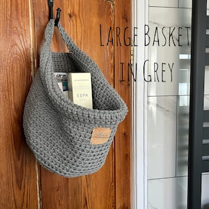 A large grey crocheted hanging basket with a brown faux leather tag that says "woolflower" hanging on a wooden door. The basket is filled with toiletries.