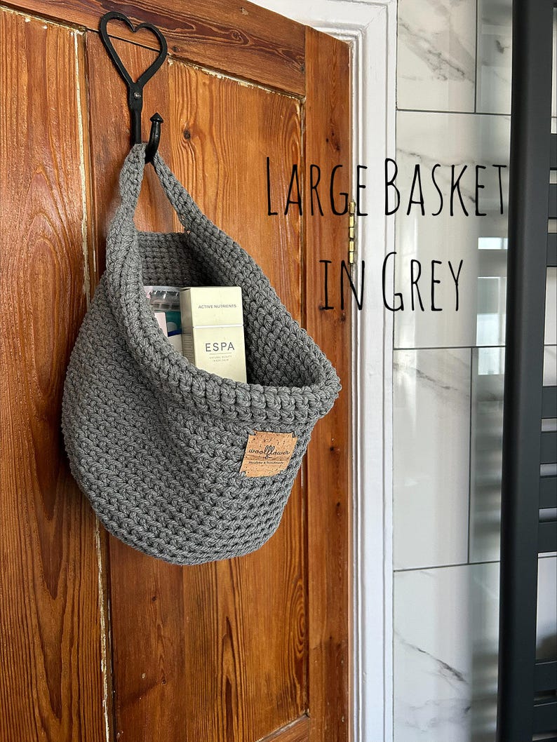 A large grey crocheted hanging basket with a brown faux leather tag that says "woolflower" hanging on a wooden door. The basket is filled with toiletries.