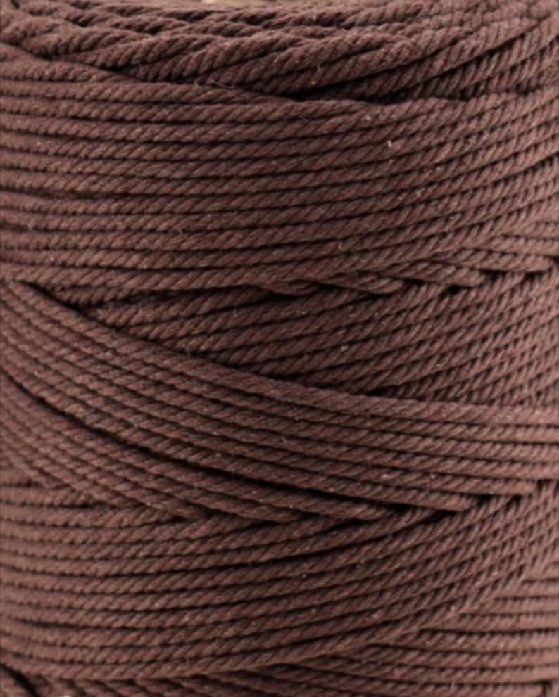 May include: A spool of dark brown macrame cord featuring a tightly twisted three-ply construction. The thick, fibrous rope is wound in uniform, horizontal layers, creating a consistent texture across the entire surface of the cylindrical spool.