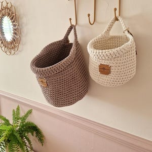 May include: Two hanging crochet baskets, one in a muted taupe and one in an off-white shade. Each basket features a single handle and a small, rectangular brown leather-like tag with the text "wildflower" embossed on the front surface.