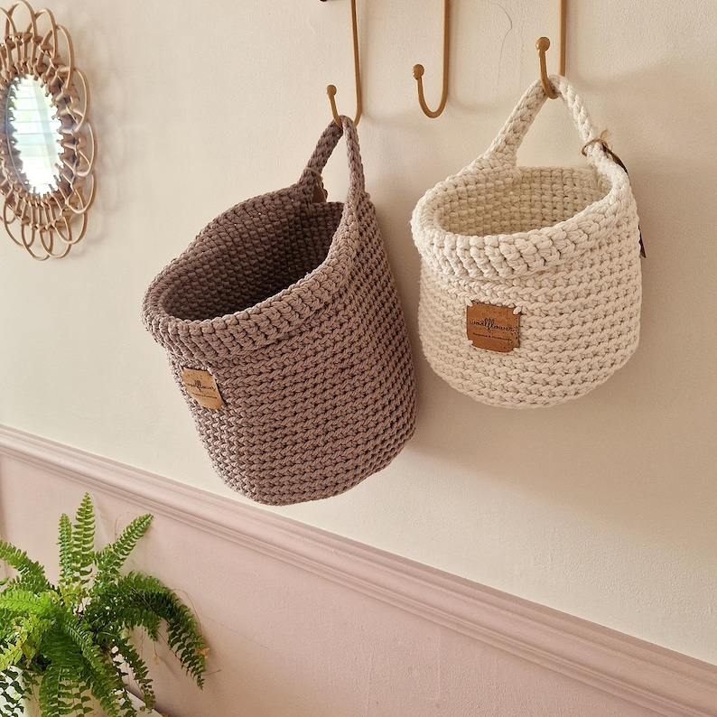 May include: Two hanging crochet baskets, one in a muted taupe and one in an off-white shade. Each basket features a single handle and a small, rectangular brown leather-like tag with the text "wildflower" embossed on the front surface.