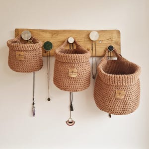 3 light brown hanging crochet baskets with a vegan friendly cork "woolflower" logo. Great for bedrooms, bathrooms and nursery home organisation on Etsy by Woolflowerdesigns!