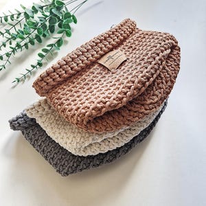 Three crocheted baskets stacked on top of each other. The top basket is slamon brown, the middle basket is off white, and the bottom basket is grey. The top basket has a brown label with the text "woolflower" on it.