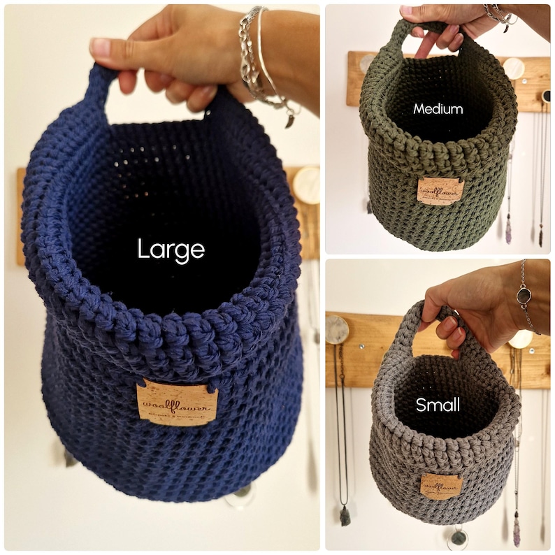 May include: Three hanging crochet baskets in navy blue, olive green, and gray. Each basket features a single handle, a textured stitch pattern, and a rectangular cork label with the text "woolflower" printed on the front. Labels indicate sizes as Large, Medium, and Small.