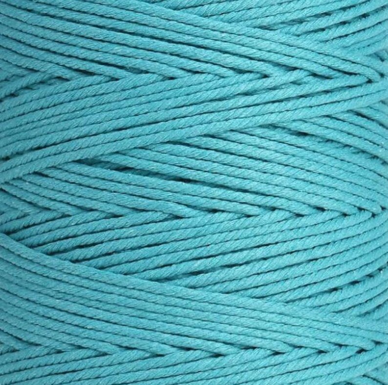 May include: A spool of turquoise blue macrame cord featuring a tightly twisted three-ply construction. The thick cotton rope is wound in a continuous, uniform pattern, creating a textured surface of diagonal lines across the entire cylindrical spool.