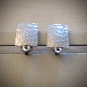 May include: Two silver-toned square cabinet knobs with a hammered texture. Each knob has a small, circular base. The knobs are mounted on a light-colored surface, suggesting they are for furniture or cabinetry.