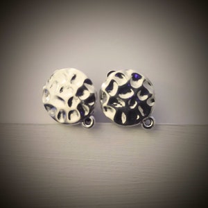 May include: A pair of silver-colored, hammered-texture earrings. Each earring is a small, round shape with a textured surface and a small loop at the bottom. The earrings are displayed against a neutral background.