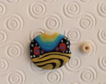 May include: A colorful glass bead with a black, yellow, blue, red, and white abstract design. The bead is oval shaped and has a glossy finish.