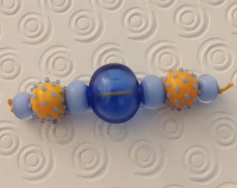 May include: A close-up of a blue and yellow beaded necklace. The necklace is made of three different types of beads: a large blue bead, a smaller blue bead, and a yellow bead with blue dots. The beads are strung on a thin string.