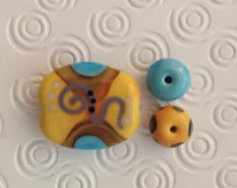 May include: Three glass beads, one with a swirl design in purple, brown, and blue on a yellow background, one solid blue, and one yellow with black dots.