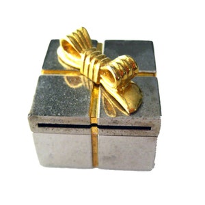 May include: A silver-colored gift box with gold-colored accents and a gold bow on top. The box is square-shaped and has a metallic finish. The gold accents are on the edges and the bow.