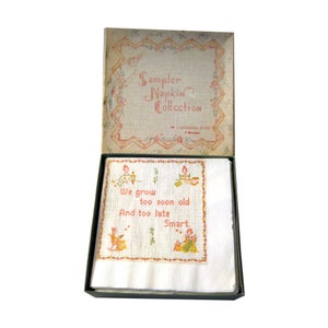 May include: A vintage sampler napkin collection box, open to reveal white napkins with embroidered designs and text. The box lid features a floral border and the words "Sampler Napkin Collection". The napkins have the text "We grow too soon old And too late Smart."