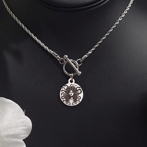 May include: A silver necklace with a toggle clasp and a round pendant featuring a detailed image of a woman's face with long hair.