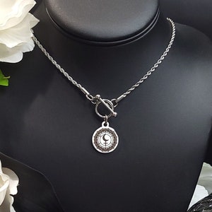 以下が含まれることがあります： 黒と白のシンボルが描かれた丸いペンダントが付いた、トグル留めのシルバーネックレス。ネックレスは、白い花が飾られた黒い背景の黒いマネキンに飾られています。