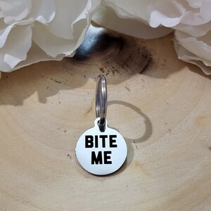 May include: A silver key chain with a round tag that says "Bite Me" in black letters.