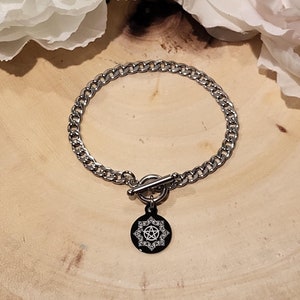 Può includere: Un braccialetto a catena argentata con un ciondolo nero che presenta un pentagramma bianco. Il braccialetto ha una chiusura a levetta.