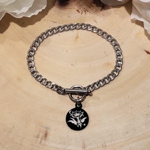 Può includere: Un braccialetto a catena argentata con un ciondolo rotondo nero con un motivo floreale bianco. Il braccialetto ha una chiusura a levetta.