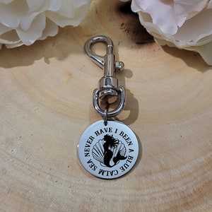 May include: Silver keychain with a round charm that says "Never have I been a blue calm sea" with a mermaid silhouette inside a seashell.