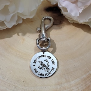 May include: A silver keychain with a round charm that says "A bird within her sang and then flew off". The charm features a small bird with a branch and leaves. The keychain is on a wooden surface with two white flowers.