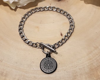 May include: A silver chain bracelet with a black metal charm featuring a geometric design. The bracelet has a toggle clasp.