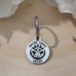 Può includere: Un portachiavi d'argento con un ciondolo rotondo che dice "Maybe Today Satan" con un diavolo dei cartoni animati con le ali e un pentagramma sulla testa.