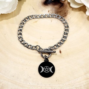 May include: A silver-colored chain bracelet with a black circular charm. The charm features a white pentagram symbol between two crescent moons. The bracelet has a toggle clasp and is displayed on a wooden surface.