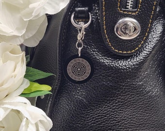 May include: Black leather handbag with a gold-colored zipper and a decorative charm. The charm is a round metal disc with a Greek key design.