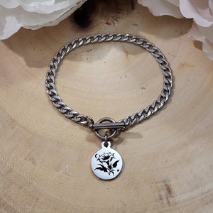 Può includere: Un braccialetto a catena d'argento con chiusura a levetta e un ciondolo rotondo con un design floreale in bianco e nero.