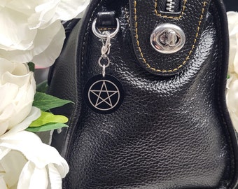 May include: Black leather handbag with a yellow stitch detail and a silver zipper. A silver charm with a black pentagram is attached to the bag.