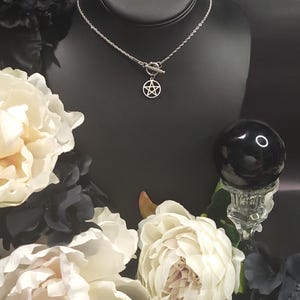 Op de afbeelding: Een zilveren ketting met een zilveren pentagram hanger en een toggle sluiting. De ketting wordt getoond op een zwarte mannequinbuste met witte bloemen op de achtergrond.
