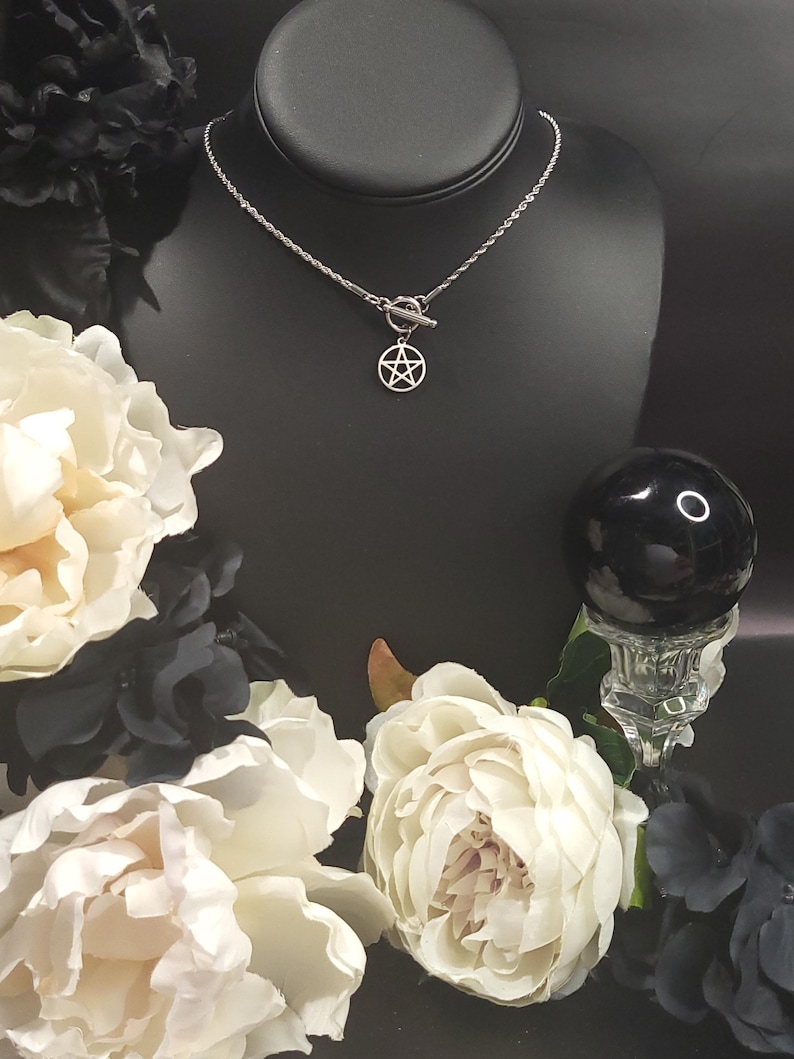 Op de afbeelding: Een zilveren ketting met een zilveren pentagram hanger en een toggle sluiting. De ketting wordt getoond op een zwarte mannequinbuste met witte bloemen op de achtergrond.