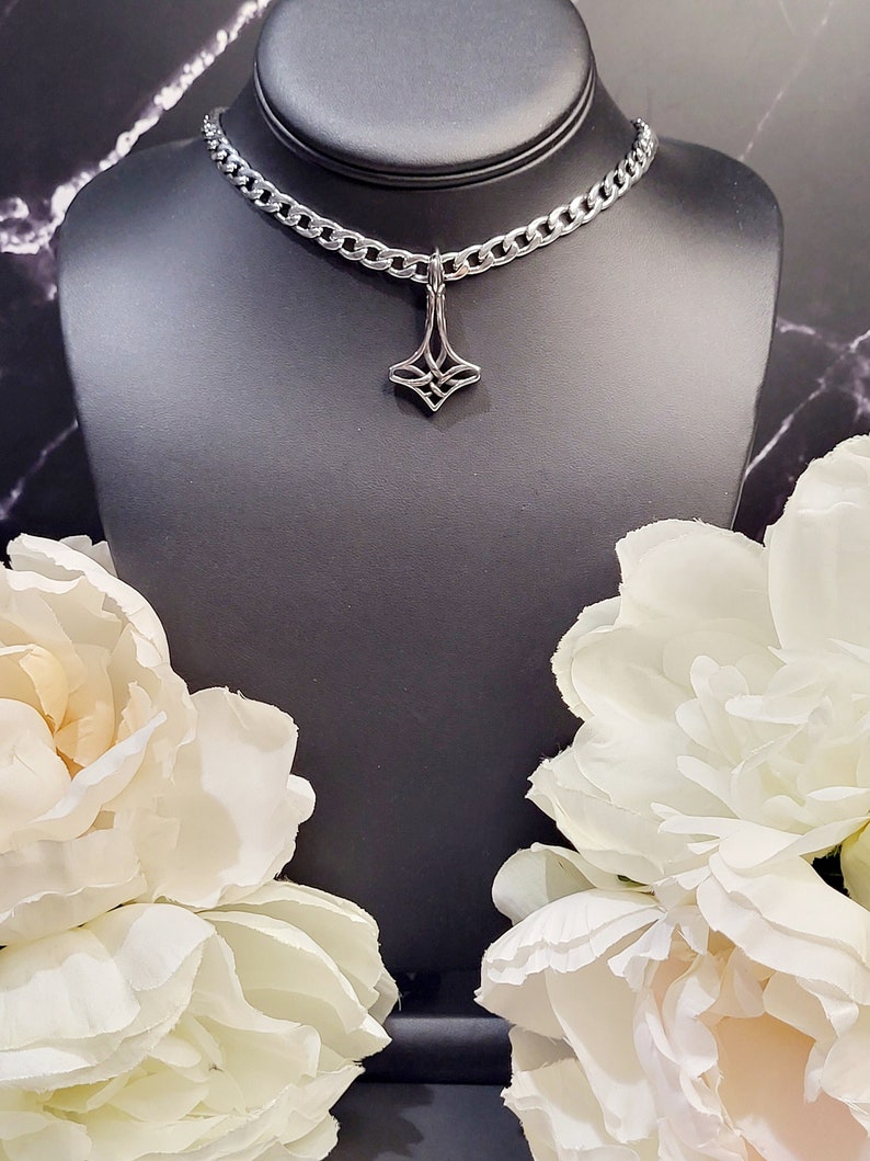 May include: A silver chain necklace with a silver pendant featuring a geometric design. The necklace is displayed on a black mannequin bust with white flowers on either side.