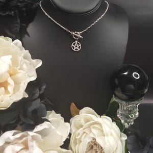 Op de afbeelding: Een zilveren ketting met een zilveren pentagram hanger en een toggle sluiting. De ketting wordt getoond op een zwarte mannequinbuste met witte bloemen op de achtergrond.