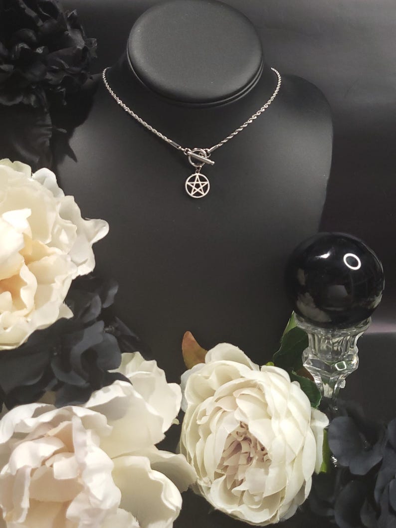 Op de afbeelding: Een zilveren ketting met een zilveren pentagram hanger en een toggle sluiting. De ketting wordt getoond op een zwarte mannequinbuste met witte bloemen op de achtergrond.
