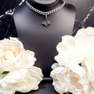 May include: A silver chain choker necklace with a geometric pendant. The necklace is displayed on a black mannequin bust with white flowers on either side.