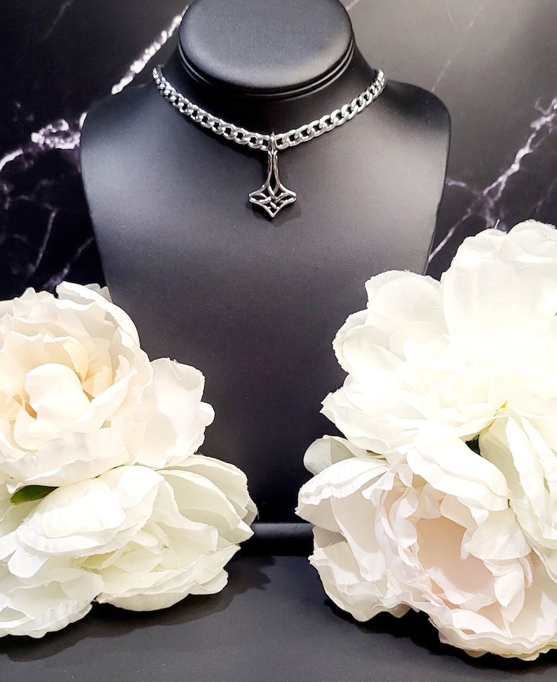 May include: A silver chain choker necklace with a geometric pendant. The necklace is displayed on a black mannequin bust with white flowers on either side.