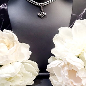 May include: A silver chain choker necklace with a geometric pendant. The necklace is on a black mannequin bust and there are white flowers on either side of the bust.